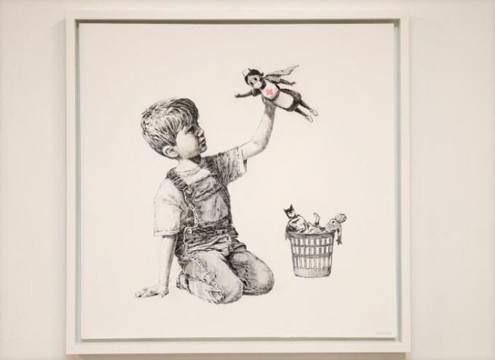 近日，Banksy 在 Instagram 上发布一幅涂鸦新作”规则改变者“（Game Changer），作品展示一名小男孩跪坐在地上玩耍的场景，他手里拿着身披英雄斗篷的护士玩偶，蜘蛛侠和蝙蝠侠则被丢进废纸篓里。据悉，这幅画作将在秋季进行拍卖，所得收入将捐赠给英国国家医疗系统。（摄影师：Stuart Martin）